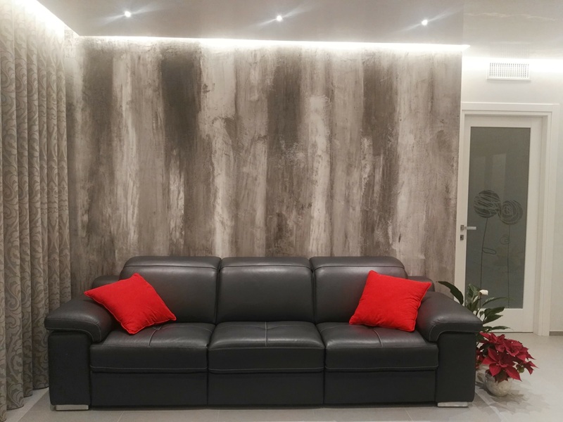 Parete decorativa grigia a striature scure sotto luce LED, divano nero con cuscini rossi, di Olive Pitturazioni
